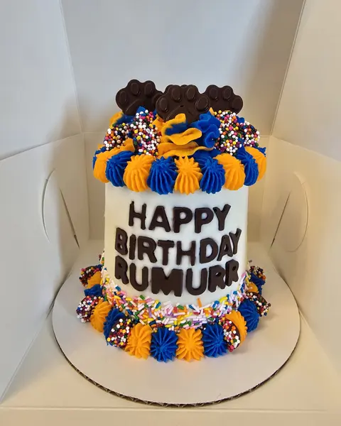 White birthday cake with HAPPY BIRTHDAY RUMURR piped on it, with blue and gold ruffles